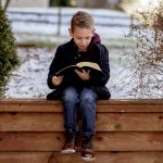 Little boy sitting on wooden planks and reading the bible in a garden covered in the snow